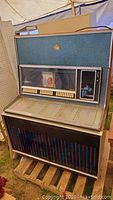Front angled view of a large vintage jukebox showcasing the upper blue textured panel with a central gold emblem, below the glass showing music selection slots and rectangular push buttons, encased in a metal frame with visible wear.