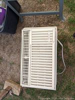 Top vent and front adjustable louvers visible on white plastic window air conditioner unit with power cord on ground outdoors.