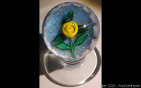 Top view showing yellow crimp rose and green leaves inside the clear glass paperweight with blue and white background and gold specks.