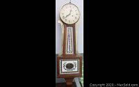 Full front view of the banjo clock, showing the round white clock face with Roman numerals and wooden body with painted floral glass panel and gold trim.