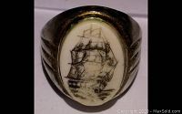 Front view of the ring showing the oval carved faux ivory stone with scrimshaw artwork of a sailing ship in black detail.