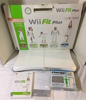 Wii Fit Plus Balance Board with original box, Wii Fit game disc and manual, and Nyko rechargeable battery pack in packaging displayed on a white cloth background.