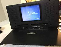 Photo of folded black video screen unit with speakers on sides, screen showing system configuration interface.