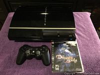 PlayStation 3 console with black wireless controller and Demon's Souls game case displayed on purple cloth