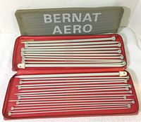 Open case containing 24 Bernat Aero grey knitting needles of various sizes organized in red velvety slots, case top reads 'BERNAT AERO'
