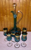 Full set of a teal colored hand blown glass wine carafe with stopper and five matching glasses, decorated with gilt and red stones, displayed on a white cloth.