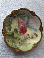 Top view of the hand painted Royal Austria plate showing embossed floral design with pink and cream thistles, signed by Jarm, and embossed gold scalloped edges.