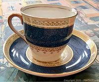 Close-up photo of demitasse cup showing cobalt blue band with gold trim and detail, white base with gold filigree, and curved handle.