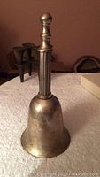 Silverplate hand bell standing upright on white cloth surface with grooved handle and rounded finial.
