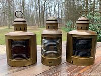 Three copper-finish ship oil lamps side by side, showing red, clear, and dark lenses against outdoor background