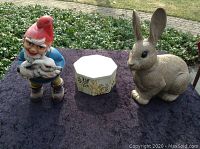 Photo of antique garden gnome holding rabbit, garden bunny figurine, and spring floral octagonal trinket box on outdoor surface