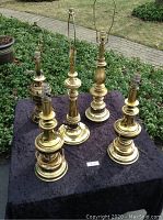 Five brass table lamps arranged on a dark cloth with garden background, showing variation in size and style, clearly displaying light sockets and bases