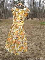 Front view of vintage floral garden party dress hanging on a hanger outdoors, showing full length and floral pattern with yellow belt.