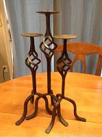 Photo of three wrought iron candle holders with twisted stems on a wooden table with a chair and curtain background.