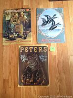 Three vintage style metal tin poster signs arranged on wooden floor showing Remington UMC, Winchester, and Peters cartridge images