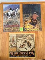 Three vintage style metal tin signs lying on wooden floor, showing hunting and cartridge advertising themes. Signs measured around 11x16 inches with folded edges and holes in corners.