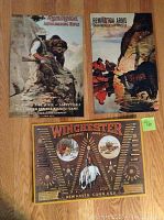 Three vintage style metal poster tin signs displayed on floor: Remington Autoloading Rifle with hunter and bear, Remington Arms with men boating by water and dog, Winchester Arms with rifles and cartridge belts on dark background.