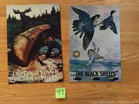 Two vintage style metal tin signs shown side by side on wooden floor. Left sign shows man in canoe and moose, right sign shows flying black ducks titled 'The Black Shells'.