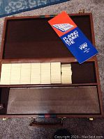 Open wooden carrying case showing organized rows of unnumbered cream-colored rummy tiles and instruction booklet on the interior lid
