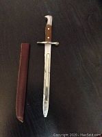 Antique bayonet with leather sheath laid on dark background, showing full length with tip broken.