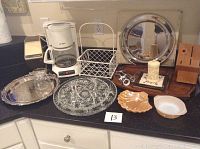 Kitchen serving pieces and appliances arranged on countertop including coffee maker, ice crusher, tray, glass trays, ramekin, wooden tray, metal basket, and utensils.