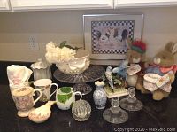 Wide view of lot items on kitchen counter including mugs, cake stand, candlesticks, plush rabbits and framed rooster print.