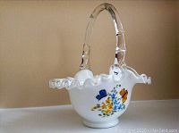 White opaque glass basket with clear textured handle and hand-painted floral patterns on the side.