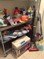Metal wire shelving with cleaning and laundry supplies including detergent bottles, cleaning sprays, and fabric softener; blue plaid ironing board beneath; coiled electrical cords and boxed items on floor