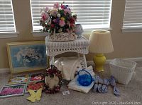 Wide view showing overall lot contents including wicker table and basket, lamp, flower arrangements, framed art, glass basket, and tablecloths