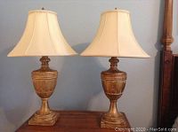Pair of two wooden table lamps with gold textured finish on a wooden surface, cream fabric bell lampshades.