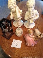 Overview of all items on a wooden table: Josef Originals music box girl at piano in pink dress, pair of saxophone busts (girl and boy), Lenox trinket dish, golden anniversary award with angel on wooden base.