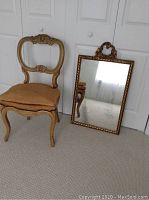 Full view of vintage carved wood chair with upholstered seat next to ornate rectangular mirror, placed against white closet doors on carpeted floor.