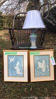 Two framed porcelain relief artworks and a table lamp displayed on a wooden chair outside