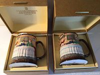 Two Royal Doulton ceramic steins displayed in original gold-toned textured boxes with cream lining. Each stein shows a painted detailed scene of people seated at a table with brown handles and bases.