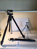 Image showing large black tripod with legs extended, small silver tripod in background, and monopod lying horizontally in front.