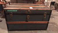 Front view of vintage storage trunk chest with dark metal body and wooden trim, featuring metal latches and central locking mechanism.