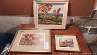 Three framed prints on wooden table against concrete wall. Top: autumn landscape. Bottom left: red foliage. Bottom right: rural carriage scene.