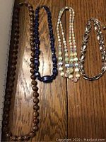 Four necklaces and a silver-tone bracelet laid out on wood surface: brown bead necklace, blue bead necklace, triple strand pastel beaded necklace, silver-tone linked bracelet with chain extender.