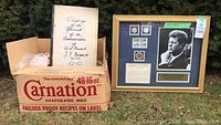Framed JFK commemorative display with coins and stamps shown alongside a cardboard box labeled Carnation evaporated milk.