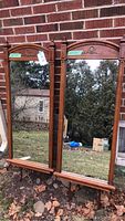 Two identical wood framed standing mirrors leaning against a brick wall outside. Each has a carved floral wreath accent in the wood crest and easel support legs visible.
