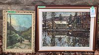 Two framed paintings leaning against a wall outdoors; one painting shows a forested path with two figures, the other a house reflected on a river.