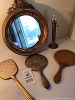 Photo showing large round ornate wall mirror, three handheld mirrors with varied styles and the brass oil lamp, all arranged together