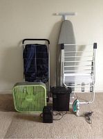Photo showing laundry cart, ironing board, drying rack, fan, space heater, paper shredder, and iron arranged on carpeted floor against wall