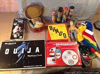 Lot contents showing vintage games and puppets including Ouija board, Master Mind, Spirograph, Bingo game, Woody figure, plush parrot puppet, and monster puppet.