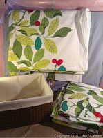 Photo of four folded linen curtains with green leaf and red berry pattern alongside the woven storage box with fabric lining.