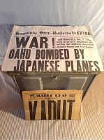 Front view of closed metal army storage box with two vintage newspapers laid on top showing distinct headlines about Pearl Harbor bombing and Maple Leaf newspaper.