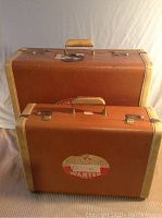 Two brown vintage travel suitcases with cream-colored edges and handles, one larger and one smaller, with travel stickers on the front.