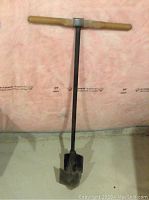 Full view of manual post hole auger with wooden handle and metal shaft ending in spiral blades.