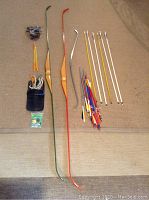 Two wooden bows (one green and one orange), one small metal bow, multiple arrows in various lengths with multicolored fletching, leather pocket holder with archery tips, package of small archery accessories laid out on carpet.