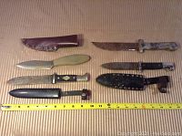 Four hunting knives laid flat next to ruler showing sizes. Knives show rust and wear on blades and handles. Two sheaths shown, one brown leather and one black leather with button strap.